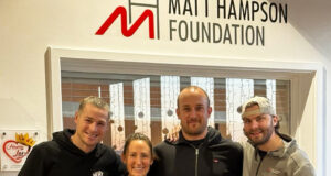 5 people pose for the camera. 4 people stand wearing running kit behind a man in a wheelchair in a coat and a blanket over his legs