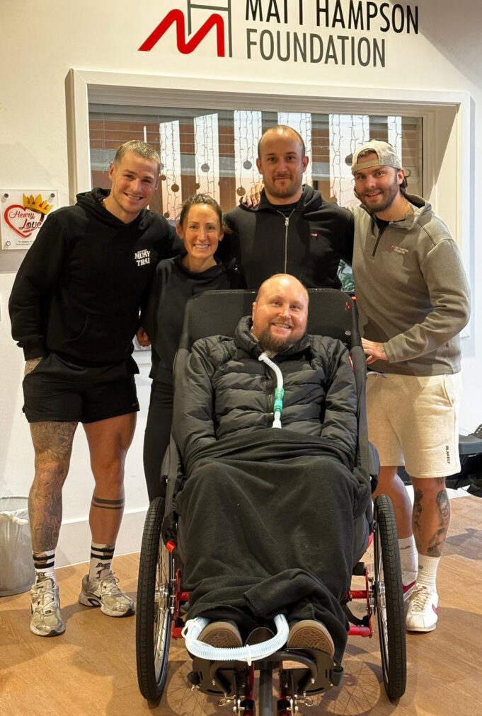 5 people pose for the camera. 4 people stand wearing running kit behind a man in a wheelchair in a coat and a blanket over his legs