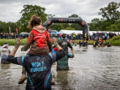 Runners wade through a waist deep body of muddy water. Some of them have children on their shoulders.