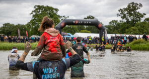 Runners wade through a waist deep body of muddy water. Some of them have children on their shoulders.