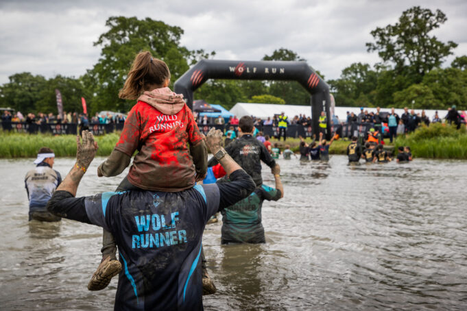 Runners wade through a waist deep body of muddy water. Some of them have children on their shoulders.