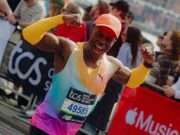 Man in brightly coloured orange/green/purple PUMA kit flexes his biceps at the camera as he runs past, smiling