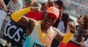 Man in brightly coloured orange/green/purple PUMA kit flexes his biceps at the camera as he runs past, smiling