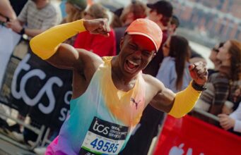 Man in brightly coloured orange/green/purple PUMA kit flexes his biceps at the camera as he runs past, smiling