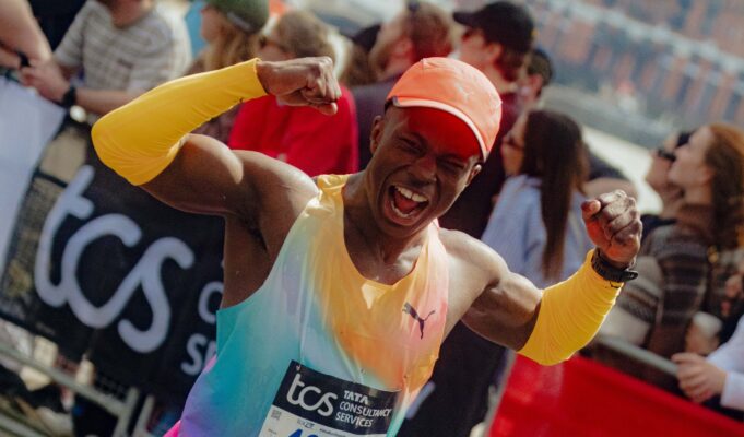 Man in brightly coloured orange/green/purple PUMA kit flexes his biceps at the camera as he runs past, smiling