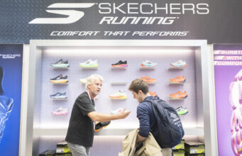 A man on a Skechers running stand shows another man some shoes