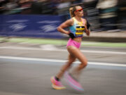 A woman with blonde hair in a ponytail and sunglasses, wearing pink shorts and a race vest, runs on a road. Image is blurred to denote fast pace.