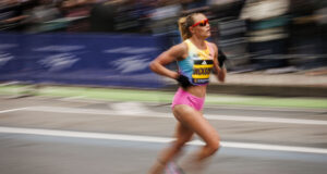 A woman with blonde hair in a ponytail and sunglasses, wearing pink shorts and a race vest, runs on a road. Image is blurred to denote fast pace.