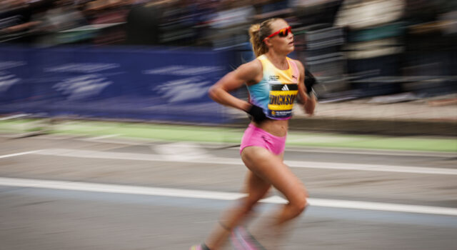 A woman with blonde hair in a ponytail and sunglasses, wearing pink shorts and a race vest, runs on a road. Image is blurred to denote fast pace.