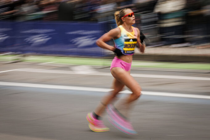 A woman with blonde hair in a ponytail and sunglasses, wearing pink shorts and a race vest, runs on a road. Image is blurred to denote fast pace.