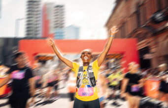A runner in yellow top put her hands iin the air in a road race. The background is blurred but there is a red gantry behind.