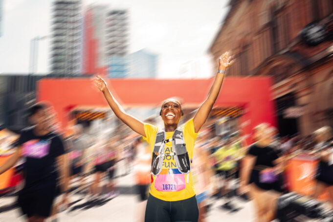 A runner in yellow top put her hands iin the air in a road race. The background is blurred but there is a red gantry behind.