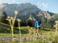 A lone male trail runner runs past craggy mountains in a green valley.