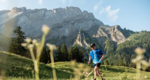 A lone male trail runner runs past craggy mountains in a green valley.