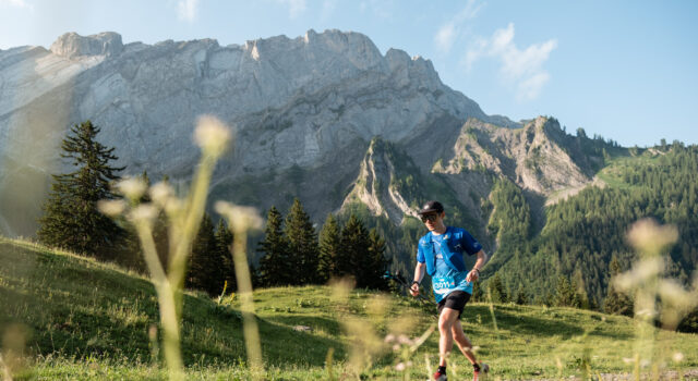 A lone male trail runner runs past craggy mountains in a green valley.