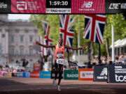Man in orange vest sprads arms wide as he runs under finish gantry of London Marathon. Buckingham Palace is visible behind him. The digital clock above his head reads 1:59:30
