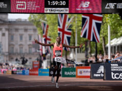 Man in orange vest sprads arms wide as he runs under finish gantry of London Marathon. Buckingham Palace is visible behind him. The digital clock above his head reads 1:59:30