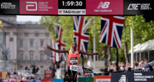 Man in orange vest sprads arms wide as he runs under finish gantry of London Marathon. Buckingham Palace is visible behind him. The digital clock above his head reads 1:59:30