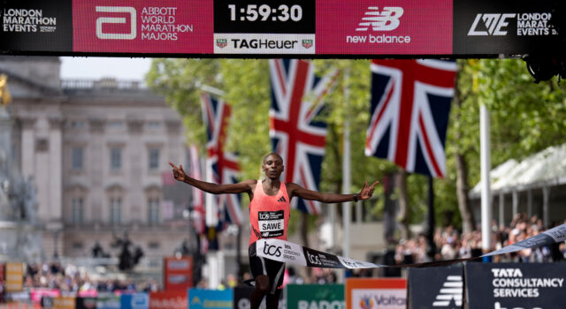 Man in orange vest sprads arms wide as he runs under finish gantry of London Marathon. Buckingham Palace is visible behind him. The digital clock above his head reads 1:59:30