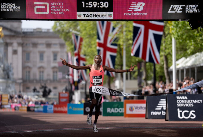 Man in orange vest sprads arms wide as he runs under finish gantry of London Marathon. Buckingham Palace is visible behind him. The digital clock above his head reads 1:59:30