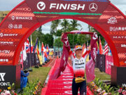 Female triathlete walks under a red finish gantry with a red finishing tape held above her head.