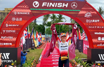 Female triathlete walks under a red finish gantry with a red finishing tape held above her head.