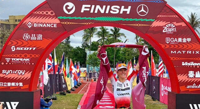 Female triathlete walks under a red finish gantry with a red finishing tape held above her head.