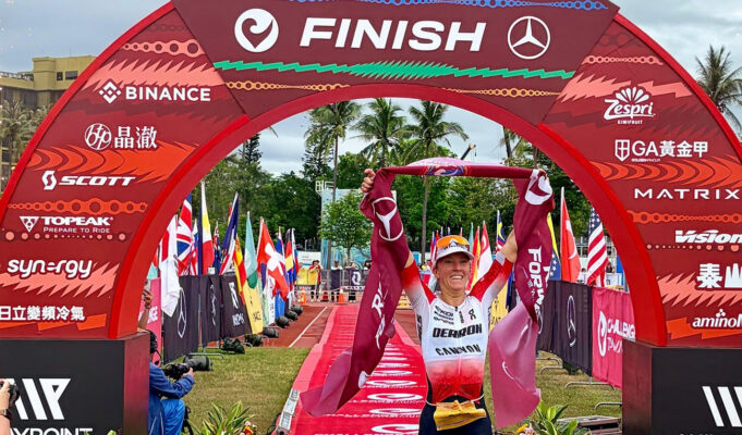 Female triathlete walks under a red finish gantry with a red finishing tape held above her head.