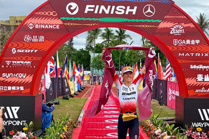 Female triathlete walks under a red finish gantry with a red finishing tape held above her head.
