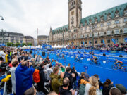 Save the date! 2027 Hamburg World Triathlon Championship Finals scheduled Spectators in front of a blue carpet in front of an ornate building. Cyclists ride past on the blue carpet.
