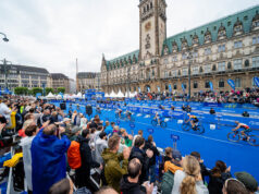 Save the date! 2027 Hamburg World Triathlon Championship Finals scheduled Spectators in front of a blue carpet in front of an ornate building. Cyclists ride past on the blue carpet.
