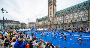 Spectators in front of a blue carpet in front of an ornate building. Cyclists ride past on the blue carpet.