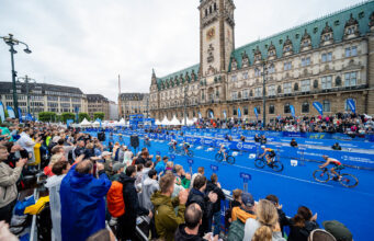 2027 Hamburg World Triathlon Championship Finals scheduled Spectators in front of a blue carpet in front of an ornate building. Cyclists ride past on the blue carpet.