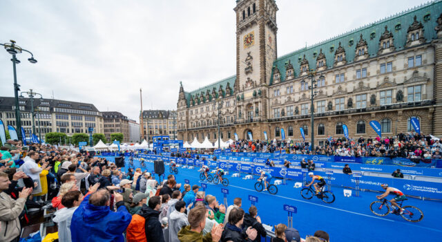 Save the date! 2027 Hamburg World Triathlon Championship Finals scheduled Spectators in front of a blue carpet in front of an ornate building. Cyclists ride past on the blue carpet.