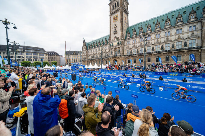 Hamburg Triathlon Spectators in front of a blue carpet in front of an ornate building. Cyclists ride past on the blue carpet.