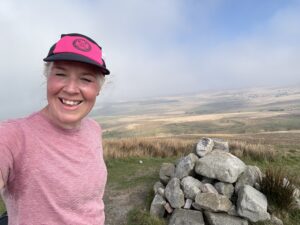 Woman in pink t-shirt and pink and black cap smiles , standing next to a cairn on a hilltop