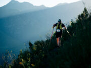 MUT by UTMB® has sold out A lone hiker on a mountainside weaves through long grass. He is wearing a race number and using nordic poles.