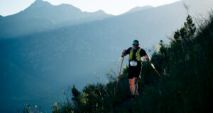 A lone hiker on a mountainside weaves through long grass. He is wearing a race number and using nordic poles.