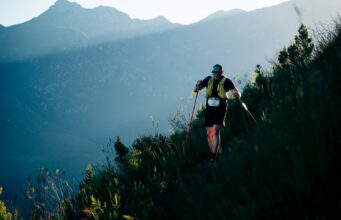 MUT by UTMB® has sold out A lone hiker on a mountainside weaves through long grass. He is wearing a race number and using nordic poles.