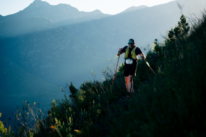 MUT-ZACZINN-48 A lone hiker on a mountainside weaves through long grass. He is wearing a race number and using nordic poles.