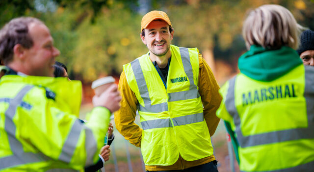 Ealing Half Marathon calls for volunteers Three marshals in hi-vis vests chat and smile. One male holds a takeaway coffee in his hand