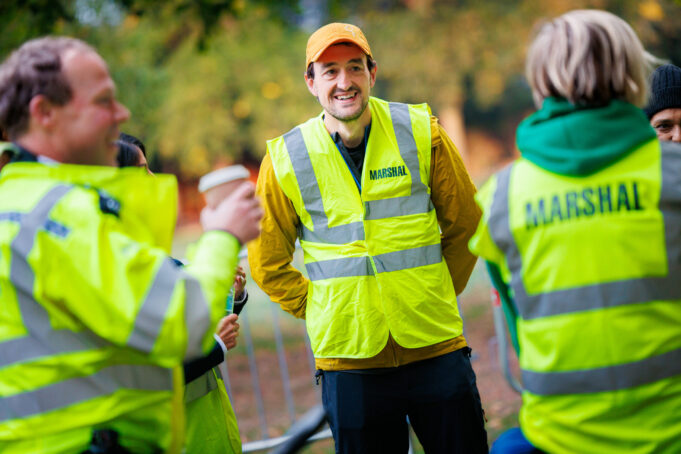 Ealing Half Volunteers Three marshals in hi-vis vests chat and smile. One male holds a takeaway coffee in his hand
