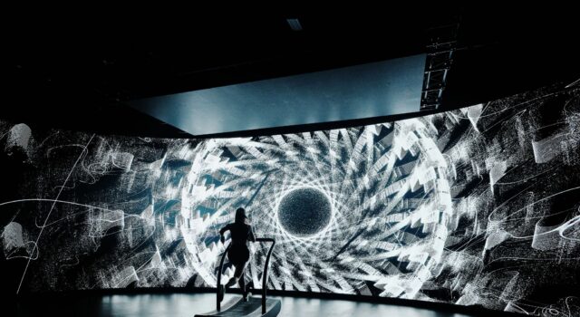 Psychedelic-inspired 24-Hour treadmill challenge A lone treadmill runner in front of a large screen showing a black and white kaleidoscopic image
