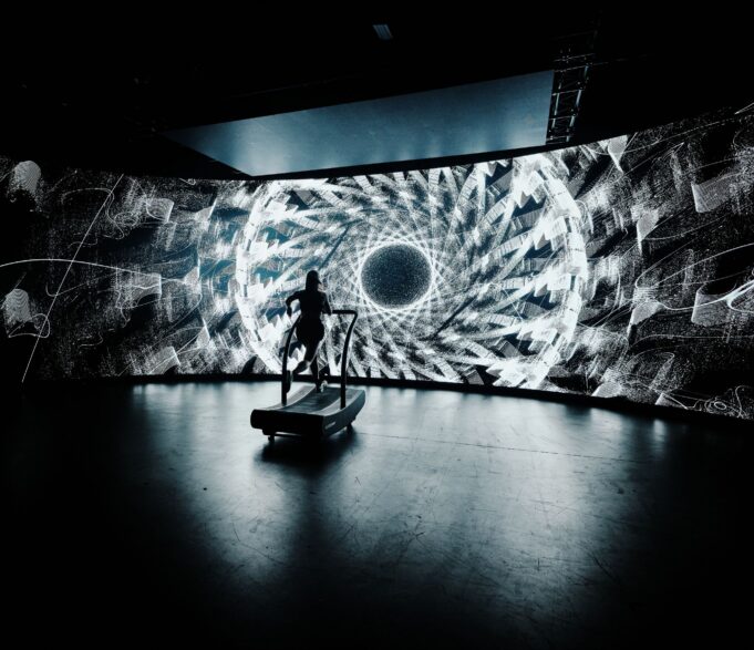 Arc Project A lone treadmill runner in front of a large screen showing a black and white kaleidoscopic image
