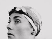 Black and whiite image of a head and shoulders shot of woman in a wetsuit with goggles and swimcap on head