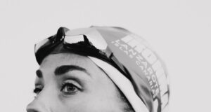 Black and whiite image of a head and shoulders shot of woman in a wetsuit with goggles and swimcap on head