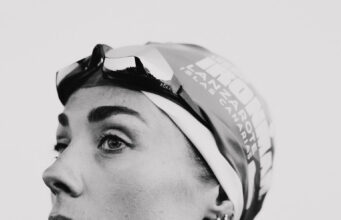 Black and whiite image of a head and shoulders shot of woman in a wetsuit with goggles and swimcap on head