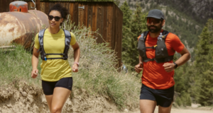 A male and female runner each wear a red and yellow t-shirt respectively, black shorts and a race vest.