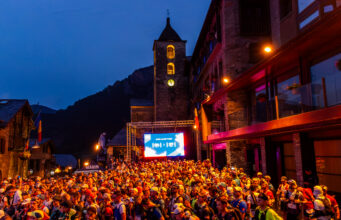 Trail 100 Andorra by UTMB® Preview A mass of runners wait at the start line at night in front of a TV screen and a lit church tower