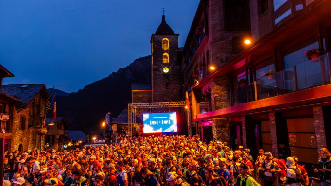 2025_Trail100 Andorra_105K DRONE @ToniGrases_0009 A mass of runners wait at the start line at night in front of a TV screen and a lit church tower